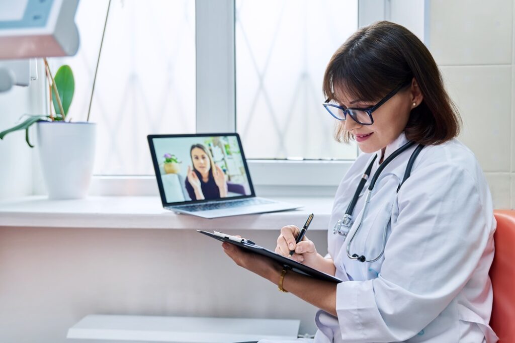 Doctor video conferencing with patient, taking notes.